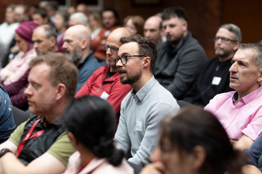 Male wearing draw glasses and a light blue shirt sitting in an audience asking a question